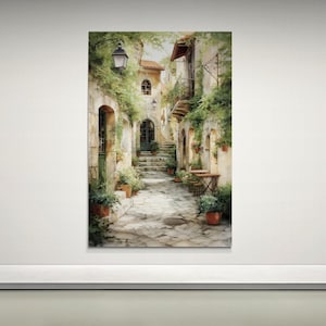Op de afbeelding: Aquarel schilderij van een Europese straatsc&egrave;ne. De afbeelding toont een smalle geplaveide straat met gebouwen, weelderig groen en potplanten. Een vintage straatlantaarn verlicht de sc&egrave;ne en kleine tafels en stoelen staan buiten.