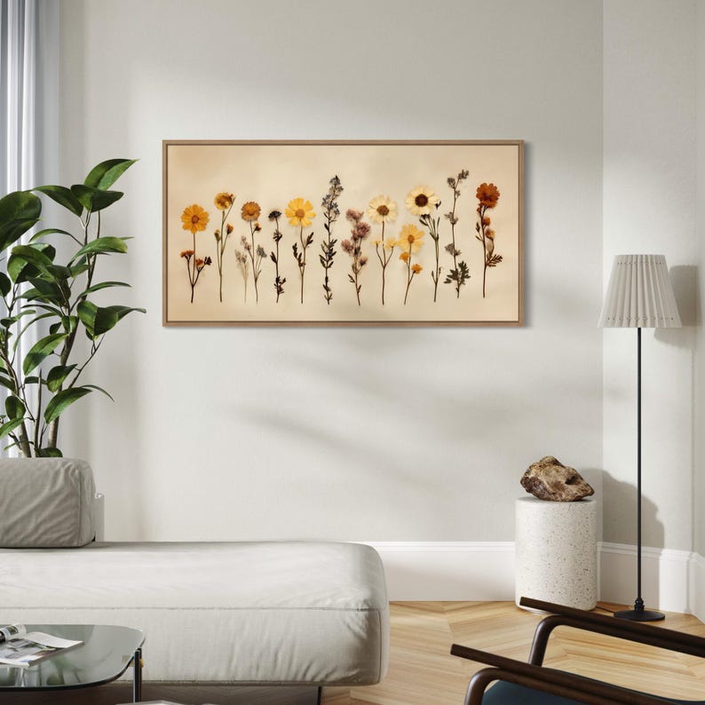May include: A framed botanical print featuring a row of dried flowers in shades of yellow, orange, and brown. The flowers are arranged against a beige background, and the frame is a light brown color. The artwork is displayed on a white wall.