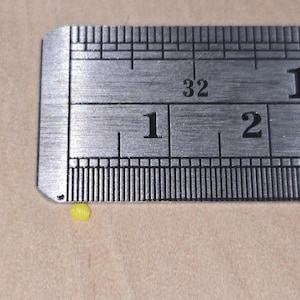 May include: A close-up of a metal ruler with black markings and numbers. The ruler shows the numbers 1 and 2, with a small yellow object near the 1-inch mark. The ruler is on a beige surface.