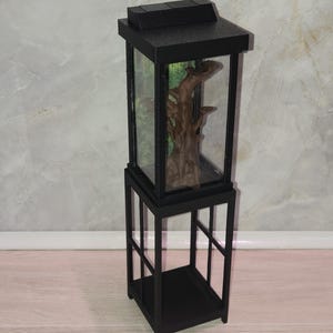 May include: A miniature black aquarium with a glass enclosure, featuring a brown driftwood decoration inside. The aquarium sits on a matching black stand. The background inside the aquarium has a green and brown forest scene.