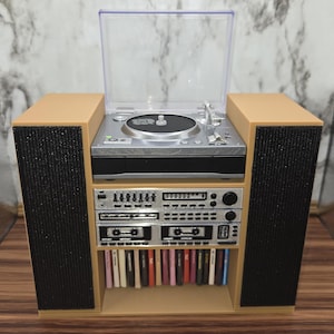 May include: A miniature stereo system featuring a silver turntable with a clear plastic dust cover, a central control unit with silver buttons and dials, two black speakers on the sides, and a lower shelf containing a row of small colorful music cassettes.