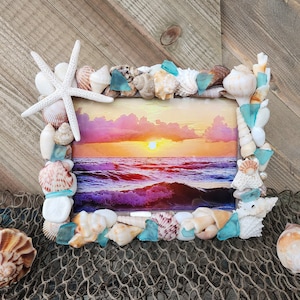 Op de afbeelding: Een fotolijst van schelpen met een zonsondergang-oceaan scène. De lijst is versierd met schelpen, zeeglas en een zeester.