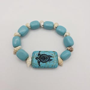 Peut inclure: Bracelet turquoise avec une pièce centrale rectangulaire ornée d'un motif de tortue noire. Le bracelet est composé de perles turquoise et de petits coquillages, pour un style estival.