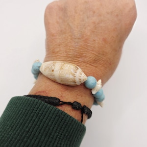 May include: A seashell bracelet featuring a large, tan seashell centerpiece and smaller white seashells and light blue beads. A black woven bracelet is also visible. The bracelet is on a wrist with a green sleeve.