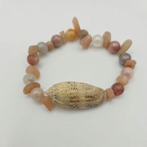 May include: A beaded bracelet featuring a natural seashell centerpiece. The bracelet is composed of various sizes and shapes of peach, tan, and gray-colored beads. The seashell is tan with a textured surface.