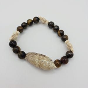 May include: A beaded bracelet featuring tiger's eye beads, small white shell accents, and a larger, oblong seashell centerpiece. The bracelet is a natural, earthy accessory with a neutral color palette.
