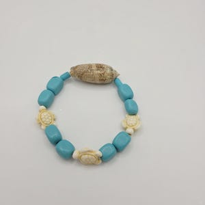 May include: A beaded bracelet featuring turquoise-colored beads, cream-colored turtle charms, and a natural seashell. The bracelet is designed with a stretchable band, perfect for easy wear. This jewelry piece is ideal for beach-themed outfits.