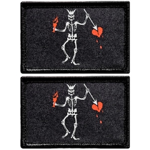 May include: Two black fabric patches with a white skeleton holding a flaming torch and a spear piercing a red heart. The patches are embroidered with white thread and have a black border.