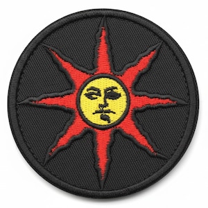 May include: A circular black patch featuring a stylized sun design. The sun has a yellow face with a stern expression, surrounded by red rays. The patch is likely an embroidered emblem, suitable for clothing or accessories.