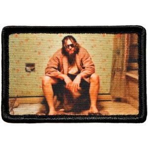 May include: Rectangular patch with a black border featuring a scene of a person in a brown robe and sunglasses sitting in a bathroom. The background shows floral wallpaper and a shower curtain.