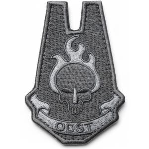 May include: A gray embroidered patch featuring a skull with flames and the text "ODST" on a banner. The patch has a unique shape with a cutout at the top. The design is in shades of gray and white.