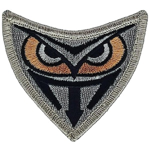 May include: A gray embroidered patch with an owl design. The owl has orange eyes and a black beak. The patch is shaped like a triangle with a pointed bottom.