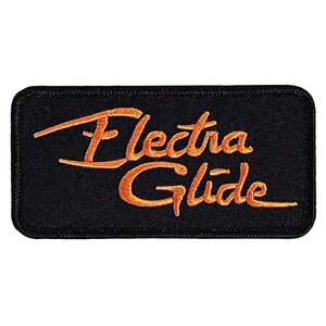 May include: Black rectangular patch with the embroidered text "Electra Glide" in orange script. The patch has a black border and is likely designed to be sewn onto clothing or accessories.
