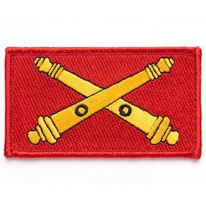 May include: A rectangular embroidered patch featuring a vibrant red background with two crossed yellow cannons. The cannons are depicted in a stylized manner with black circular details along their barrels and textured gold-toned thread.