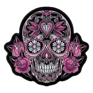 May include: Pink and white sugar skull with a diamond in the forehead and roses on the sides. The skull is embroidered on a black background.