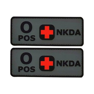 May include: Two gray rectangular patches with black stitching around the edges. Each patch has a black circle with the letter "O" inside, the words "POS" below, a red cross, and the text "NKDA" in black.