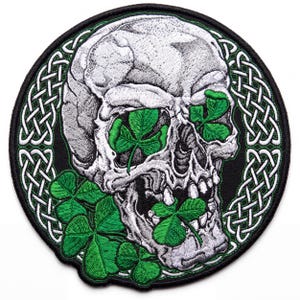 May include: A circular embroidered patch featuring a grey and white skull with bright green four-leaf clovers in its eye sockets and mouth. The skull is centered against a black background surrounded by a white and green intricate Celtic knot border.
