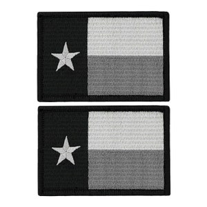 May include: Two black and white embroidered Texas state flag patches. The patches are rectangular and have a white star in the top left corner. The patches are made of a textured fabric.