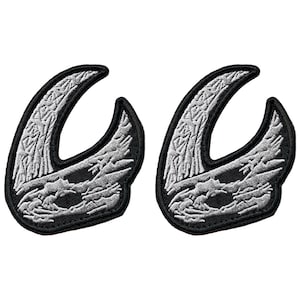 May include: Two black and white embroidered patches. Each patch features a stylized, curved design with a detailed, textured pattern in white against a black background. The patches are likely for clothing or accessories.