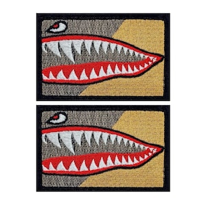 May include: Two embroidered patches featuring a stylized shark mouth design. Each patch has a black border, a tan and brown background, a red mouth, white teeth, and a red eye. The patches are rectangular.