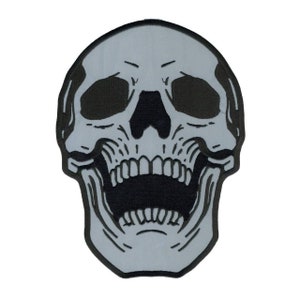 May include: A gray and black embroidered skull patch with an open mouth and teeth. The skull is outlined in black and has a detailed design.