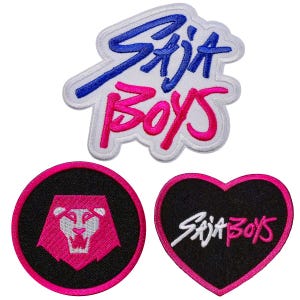May include: Embroidered patches with the text "SAJA BOYS" in blue and pink, and a lion graphic. The patches are in various shapes, including a heart and a circle, with a black and pink color scheme.