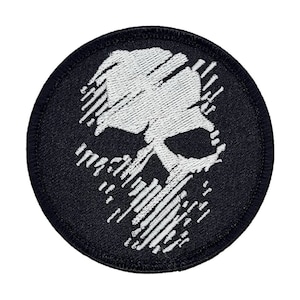 May include: A round, black embroidered patch featuring a white skull design. The skull is stylized with a distressed, hatched pattern. The patch has a black border and is likely for clothing or accessories.