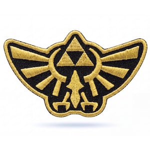 May include: An embroidered patch featuring a gold-colored Triforce symbol centered above a stylized bird crest. The design is rendered in metallic gold thread against a solid black fabric background, with a thick gold border outlining the shape.