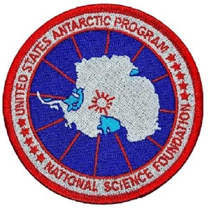 May include: A blue and red circular patch with the text "United States Antarctic Program National Science Foundation" and five stars around the outer edge. The center of the patch features a white outline of Antarctica with a red sun and blue water.