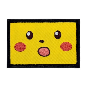 May include: A yellow embroidered patch with a cartoon face. The face has large brown eyes, a small black nose, and a red open mouth. The face is surrounded by a black border.