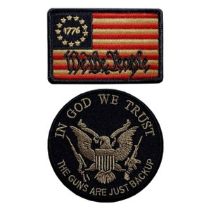 May include: Two embroidered patches. The first patch is a red, white, and gold American flag with the words "We the People" and "1776" embroidered on it. The second patch is black with a gold eagle and the words "In God We Trust" and "The Guns Are Just Backup" embroidered on it.