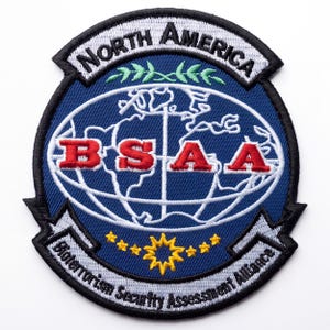 May include: A circular embroidered patch featuring a blue background with a white globe outline. The text NORTH AMERICA is at the top, BSAA in red letters across the center, and Bioterrorism Security Assessment Alliance at the bottom with yellow stars.