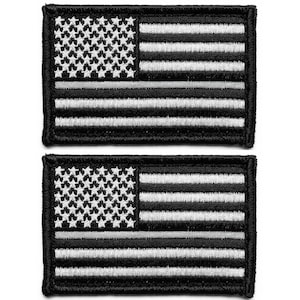May include: Two embroidered patches featuring the American flag in black, white, and gray. The flag design includes a field of white stars on a black background, white and black stripes, and a gray stripe. The patches have a black border.