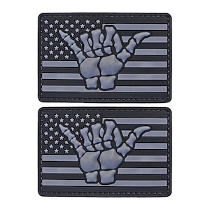 May include: Two black and white patches with a skeleton hand making the shaka sign in front of an American flag. The patches are made of a textured material.