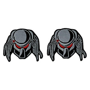 May include: Two gray and black embroidered iron-on patches of a Predator alien head with red eyes.