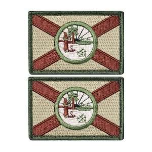 May include: Two embroidered patches featuring the state flag of Florida. The patches are rectangular and have a green border. The flag is beige with a red diagonal cross and a green circle in the center. The circle contains a depiction of a tree and a sun.