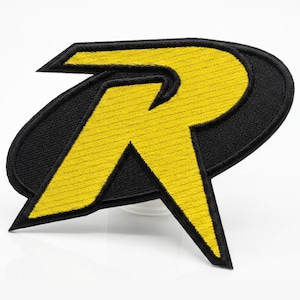 May include: A yellow and black embroidered patch featuring a stylized letter R. The design consists of a bold yellow R shape with sharp, angular edges, set against a solid black oval background with a thick black stitched border throughout.