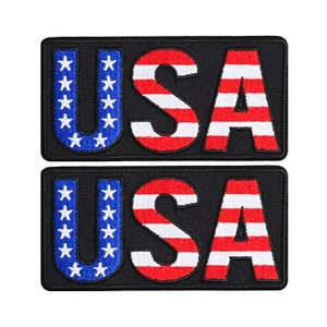 May include: Two black fabric patches with the word "USA" embroidered in white, red, and blue. The letters are outlined in black and filled with the colors of the American flag.