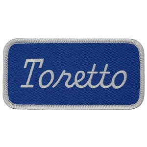 May include: A rectangular blue patch with the name "Toretto" embroidered in white script. The patch has a white stitched border, and the overall design is simple and clean.