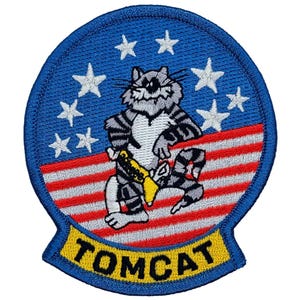 May include: A blue and yellow embroidered patch with a cartoon cat wearing a yellow belt and a red, white, and blue American flag in the background. The text "TOMCAT" is written in yellow on a blue background.