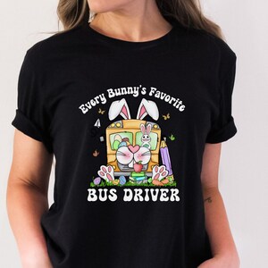 May include: Black t-shirt featuring the text "Every Bunny's Favourite Bus Driver" and a colourful graphic of a bunny driving a school bus. The graphic includes Easter eggs, butterflies, and carrots. The shirt is a casual style.