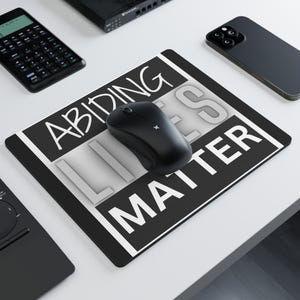 May include: Black rectangular mousepad with the text "ABIDING LIES MATTER" in white. A black wireless mouse sits on the mousepad. Other black electronics and a smartphone are visible in the background.