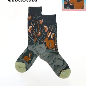May include: A pair of crew socks featuring a dark charcoal background with a pattern of brown rabbits, light pink and orange flowers, and green foliage. The lower portion of the socks displays a muted green color with wavy line details and light green toes.