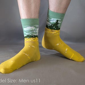 May include: A pair of crew socks featuring a landscape design with a solid mustard yellow foot and a light green cuff. The upper section displays a graphic of a dark green pine tree against a light blue sky with white clouds. Model size: Men US 11.