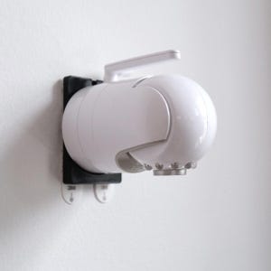 May include: A white, spherical security camera with a handle and a silver-colored base, mounted on a black bracket. The camera is attached to a white wall with two adhesive hooks.