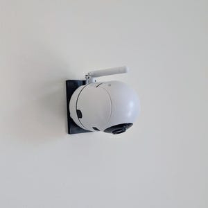 May include: A white security camera with a black base and a small antenna mounted on a white wall.