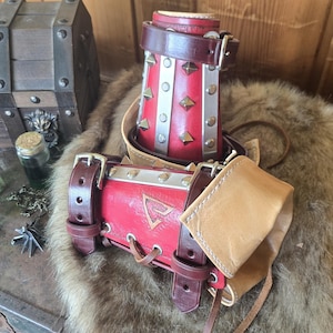Puede incluir: Un protector de brazo de cuero rojo con detalles plateados y correas marrones, junto a un recipiente de cuero rojo a juego. También se ven un cofre del tesoro de madera y pequeñas botellas de vidrio con líquido verde.