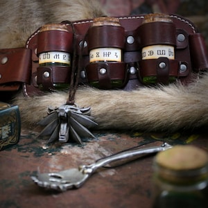 Może przedstawiać: Brązowy skórzany pasek z trzema małymi szklanymi butelkami przymocowanymi do niego. Butelki mają korkowe nakrętki i etykiety z tekstem "2 MP", "8 HP" i "20 MP". Srebrny wisiorek z motywem wilczej głowy zwisa z paska.