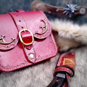 Op de afbeelding: Een rode leren tas met een gouden gesp en decoratieve metalen accenten. De tas heeft een klep met een gestikt ontwerp en ligt op een harige ondergrond.