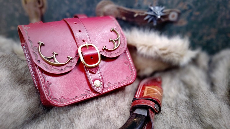 Op de afbeelding: Een rode leren tas met een gouden gesp en decoratieve metalen accenten. De tas heeft een klep met een gestikt ontwerp en ligt op een harige ondergrond.